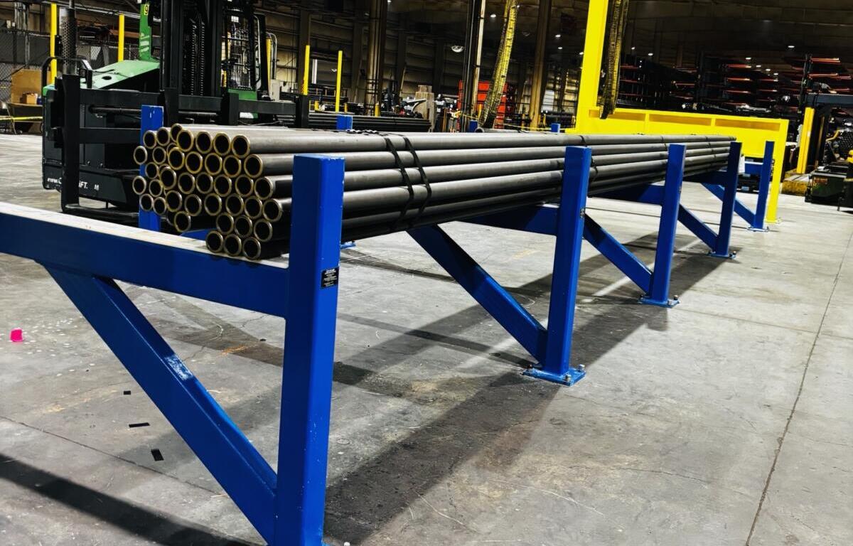 Blue industrial rack holding bundled steel pipes in a factory warehouse, with a forklift and yellow support columns in the background