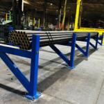Blue industrial rack holding bundled steel pipes in a factory warehouse, with a forklift and yellow support columns in the background