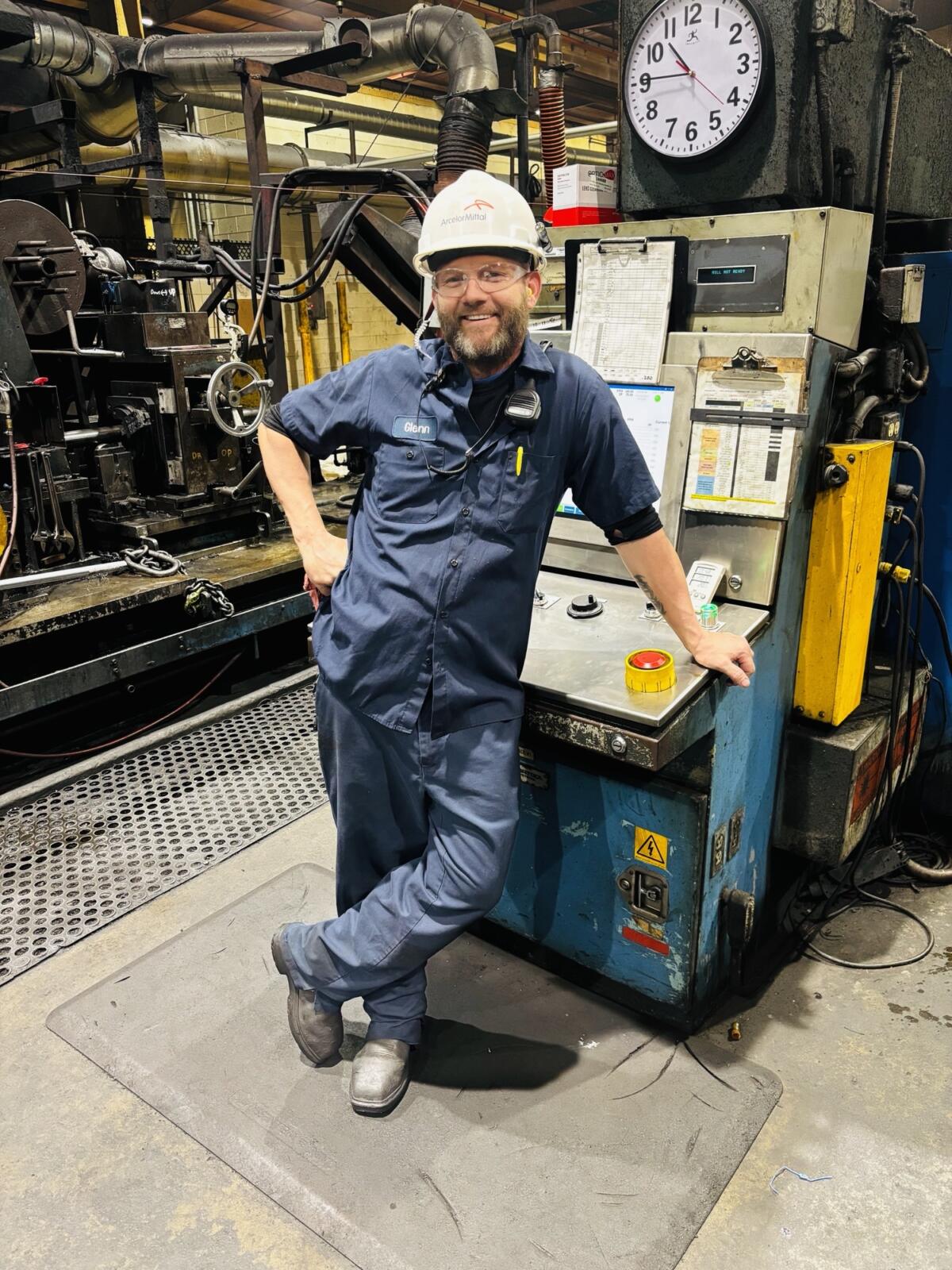 Male maintenance worker in blue coveralls, hard hat, and safety glasses leaning on a control panel beside a large industrial machine; a round clock is visible above him.