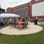 Outdoor community event with people gathered around wooden picnic tables on a circular concrete pad, beside a red brick building with arched windows and decorative theater masks.