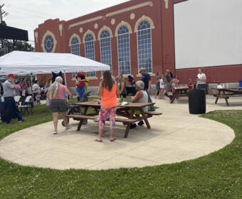 Outdoor community event with people gathered around wooden picnic tables on a circular concrete pad, beside a red brick building with arched windows and decorative theater masks.