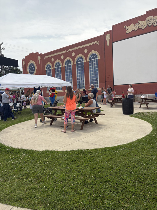 Outdoor community event with people gathered around wooden picnic tables on a circular concrete pad, beside a red brick building with arched windows and decorative theater masks.