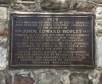 Bronze commemorative plaque mounted on a rough stone wall honoring John Edward Hopley and his contributions to Lincoln Highway, dated 1929 with biographical details etched in gold lettering.