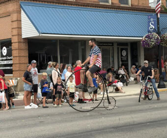 Man on a tall unicycle rides along a street parade while bystanders watch outside brick storefronts with a blue awning.