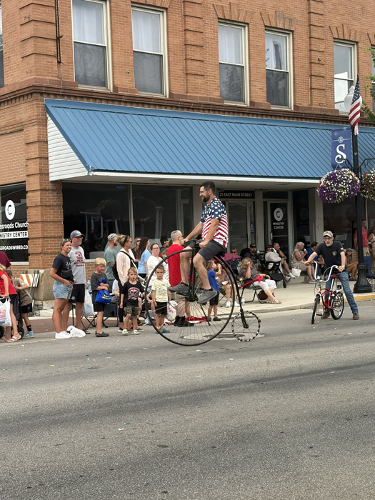 Man on a tall unicycle rides along a street parade while bystanders watch outside brick storefronts with a blue awning.
