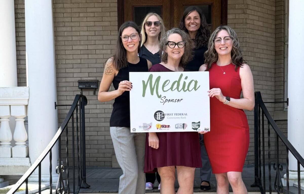 Six women standing on porch steps, holding a white 'Media Sponsor' sign and smiling at the camera.