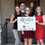 Six women standing on porch steps, holding a white 'Media Sponsor' sign and smiling at the camera.
