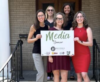 Six women standing on porch steps, holding a white 'Media Sponsor' sign and smiling at the camera.
