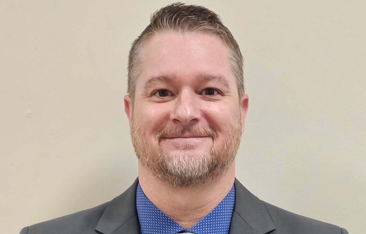Smiling man in a gray suit jacket and blue patterned shirt against a beige wall, head-and-shoulders portrait.