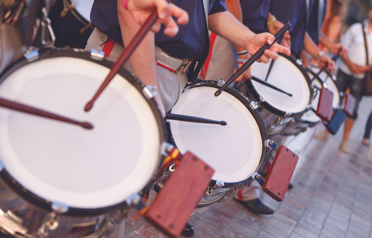 Row of marching drummers playing large white-banded bass drums in a parade, sticks mid-air as they beat in rhythm.