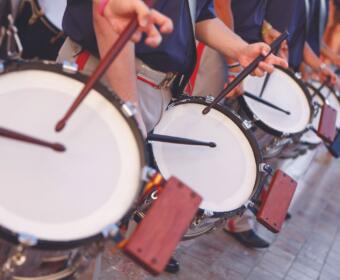 Row of marching drummers playing large white-banded bass drums in a parade, sticks mid-air as they beat in rhythm.