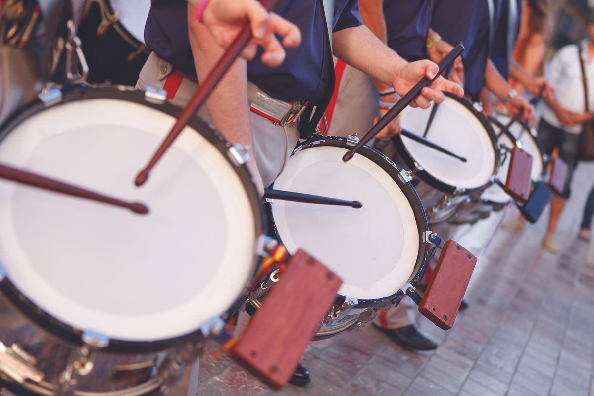 Row of marching drummers playing large white-banded bass drums in a parade, sticks mid-air as they beat in rhythm.