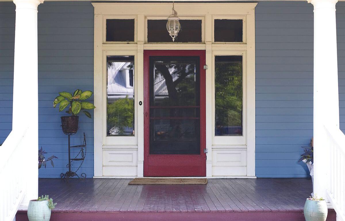 Front porch of a blue house with a red screen door, white trim, and potted plants on the steps.