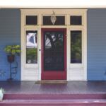 Front porch of a blue house with a red screen door, white trim, and potted plants on the steps.