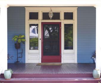 Front porch of a blue house with a red screen door, white trim, and potted plants on the steps.