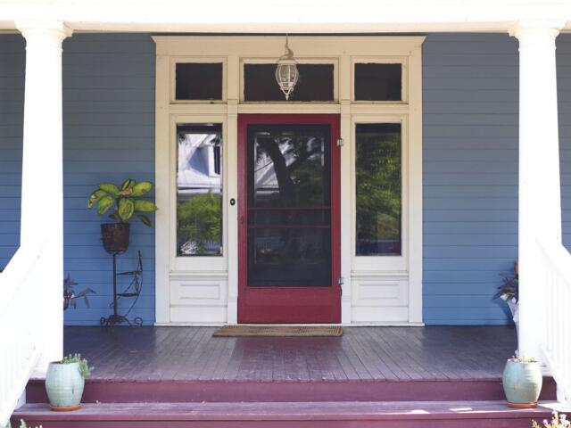 Front porch of a blue house with a red screen door, white trim, and potted plants on the steps.