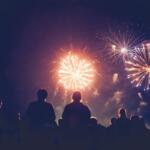 Silhouetted crowd watching bright fireworks explode in a dark night sky.
