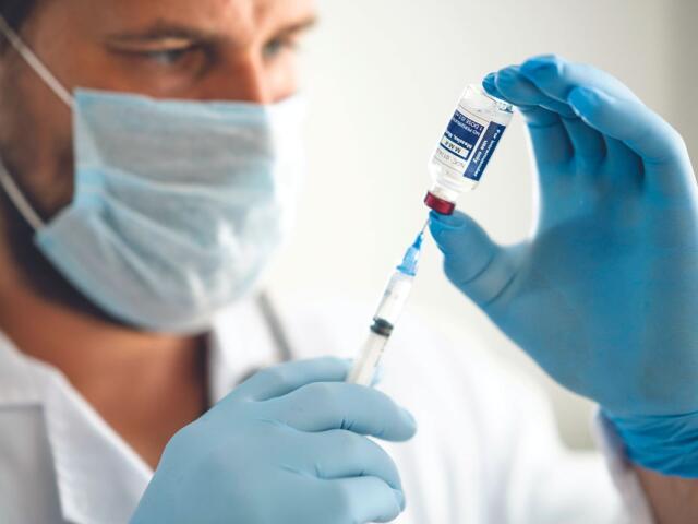 Healthcare worker in mask and blue gloves draws vaccine from a vial into a syringe text to administer a shot