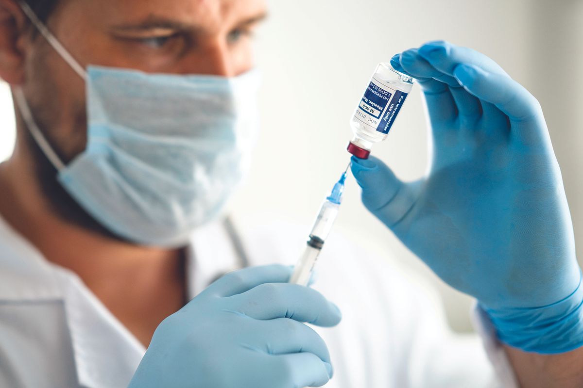 Healthcare worker in mask and blue gloves draws vaccine from a vial into a syringe text to administer a shot