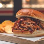 Pulled pork sandwich with BBQ sauce on a soft bun, served with potato chips on the side.