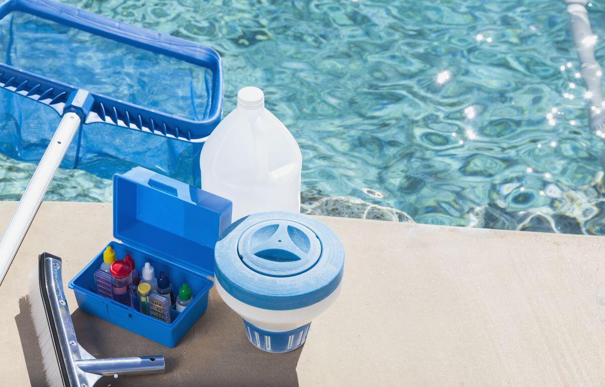 Open blue toolbox with pool chemicals and dispensing bottles on a concrete pool deck beside a pool skimmer/float adapter and water in the background