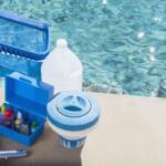 Open blue toolbox with pool chemicals and dispensing bottles on a concrete pool deck beside a pool skimmer/float adapter and water in the background