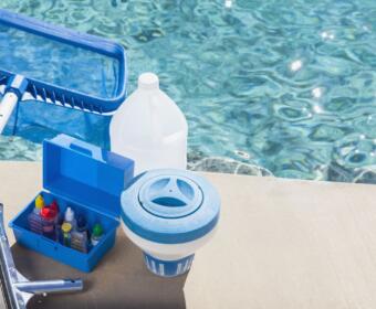 Open blue toolbox with pool chemicals and dispensing bottles on a concrete pool deck beside a pool skimmer/float adapter and water in the background