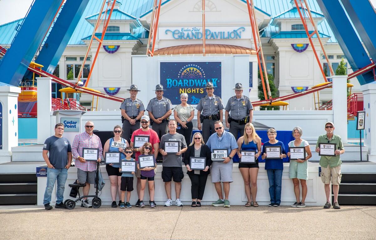 Group photo on the Cedar Point Boardwalk Nights stage in front of the Grand Pavilion: adults and children holding certificates, with uniformed officers lined up behind them, outdoors on a sunny day.