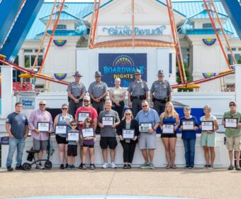 Group photo on the Cedar Point Boardwalk Nights stage in front of the Grand Pavilion: adults and children holding certificates, with uniformed officers lined up behind them, outdoors on a sunny day.