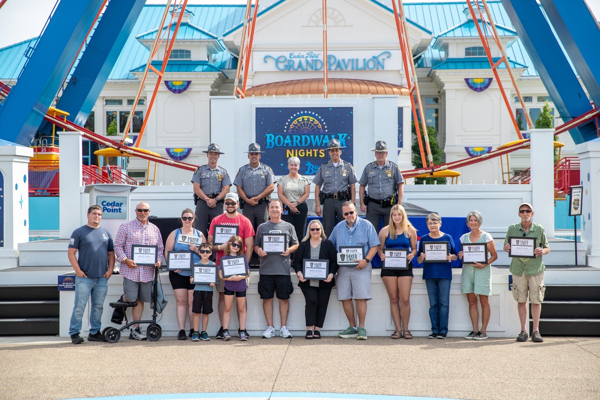 Group photo on the Cedar Point Boardwalk Nights stage in front of the Grand Pavilion: adults and children holding certificates, with uniformed officers lined up behind them, outdoors on a sunny day.