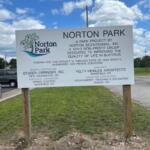 Norton Park sign listing park project sponsors and contractors, mounted on two wooden posts in a grassy lot.