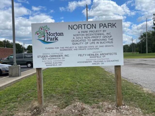 Norton Park sign listing park project sponsors and contractors, mounted on two wooden posts in a grassy lot.