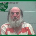Mugshot of an older man with a long gray beard in an orange-and-white striped jumpsuit, against a gray cinder-block wall; Crawford County Now logo in the upper left.