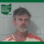 Mugshot of a middle-aged man with gray hair and stubble in an orange-and-white striped jail jumpsuit, standing against a gray cinder block wall; Crawford County Now logo in the upper left corner.