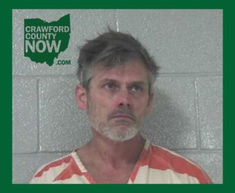 Mugshot of a middle-aged man with gray hair and stubble in an orange-and-white striped jail jumpsuit, standing against a gray cinder block wall; Crawford County Now logo in the upper left corner.
