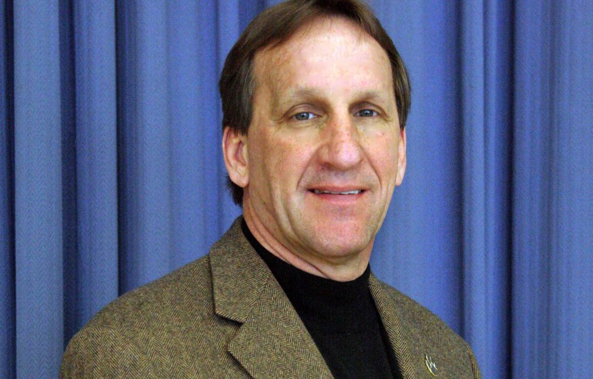 Head-and-shoulders portrait of a man in a brown tweed blazer against a blue curtain backdrop.