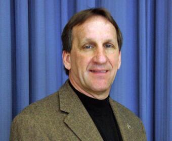 Head-and-shoulders portrait of a man in a brown tweed blazer against a blue curtain backdrop.