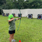 Girl in a green cap shoots a bow at outdoor archery targets on a grassy field, with an orange cone nearby.