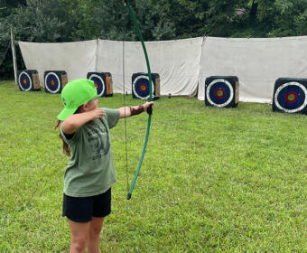 Girl in a green cap shoots a bow at outdoor archery targets on a grassy field, with an orange cone nearby.