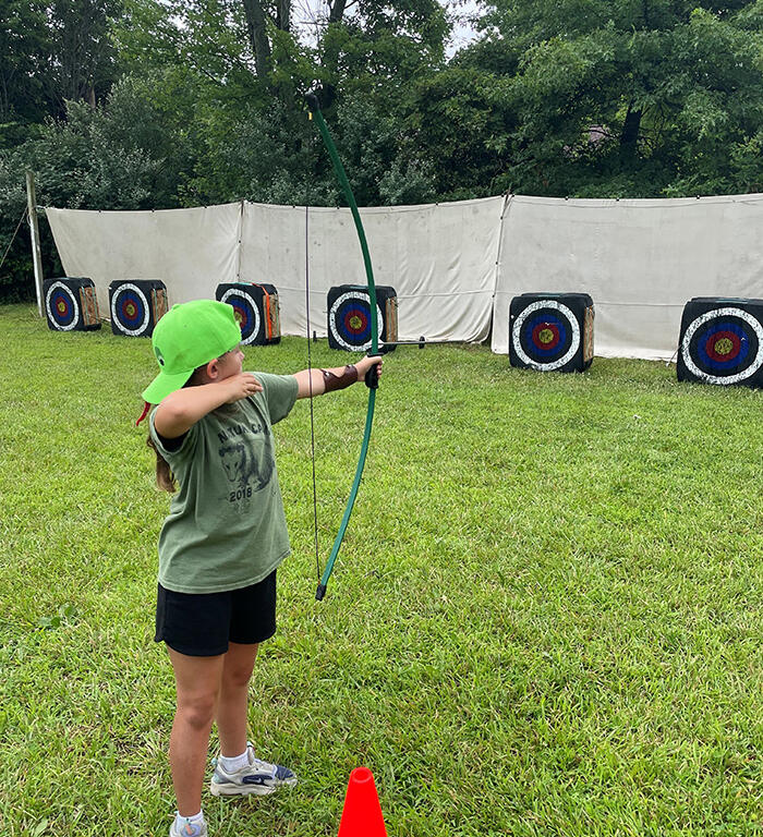 Girl in a green cap shoots a bow at outdoor archery targets on a grassy field, with an orange cone nearby.
