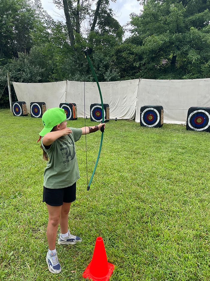 Girl in a green cap shoots a bow at outdoor archery targets on a grassy field, with an orange cone nearby.