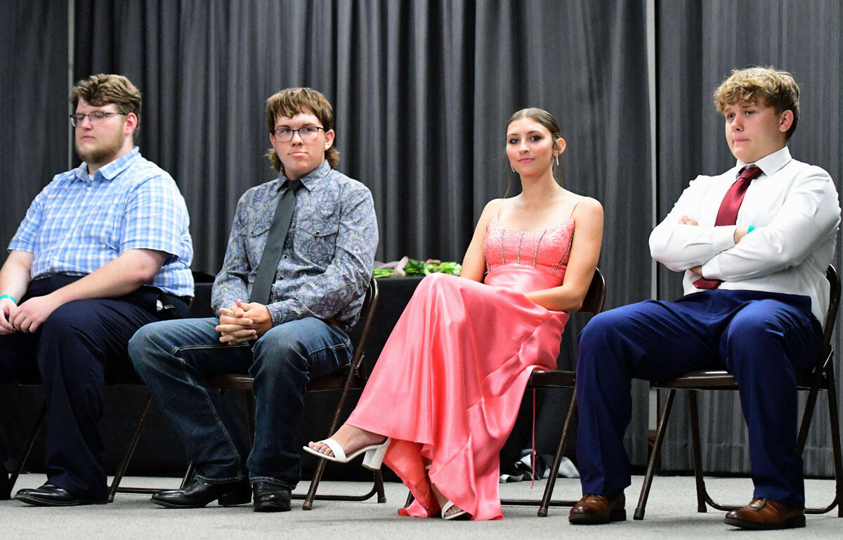 Four young people sit in a row on a stage, dressed in formal attire, facing forward against a dark curtain backdrop.