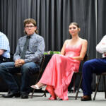 Four young people sit in a row on a stage, dressed in formal attire, facing forward against a dark curtain backdrop.