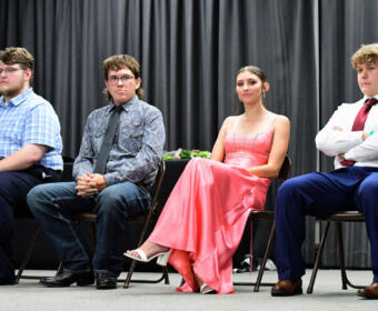 Four young people sit in a row on a stage, dressed in formal attire, facing forward against a dark curtain backdrop.