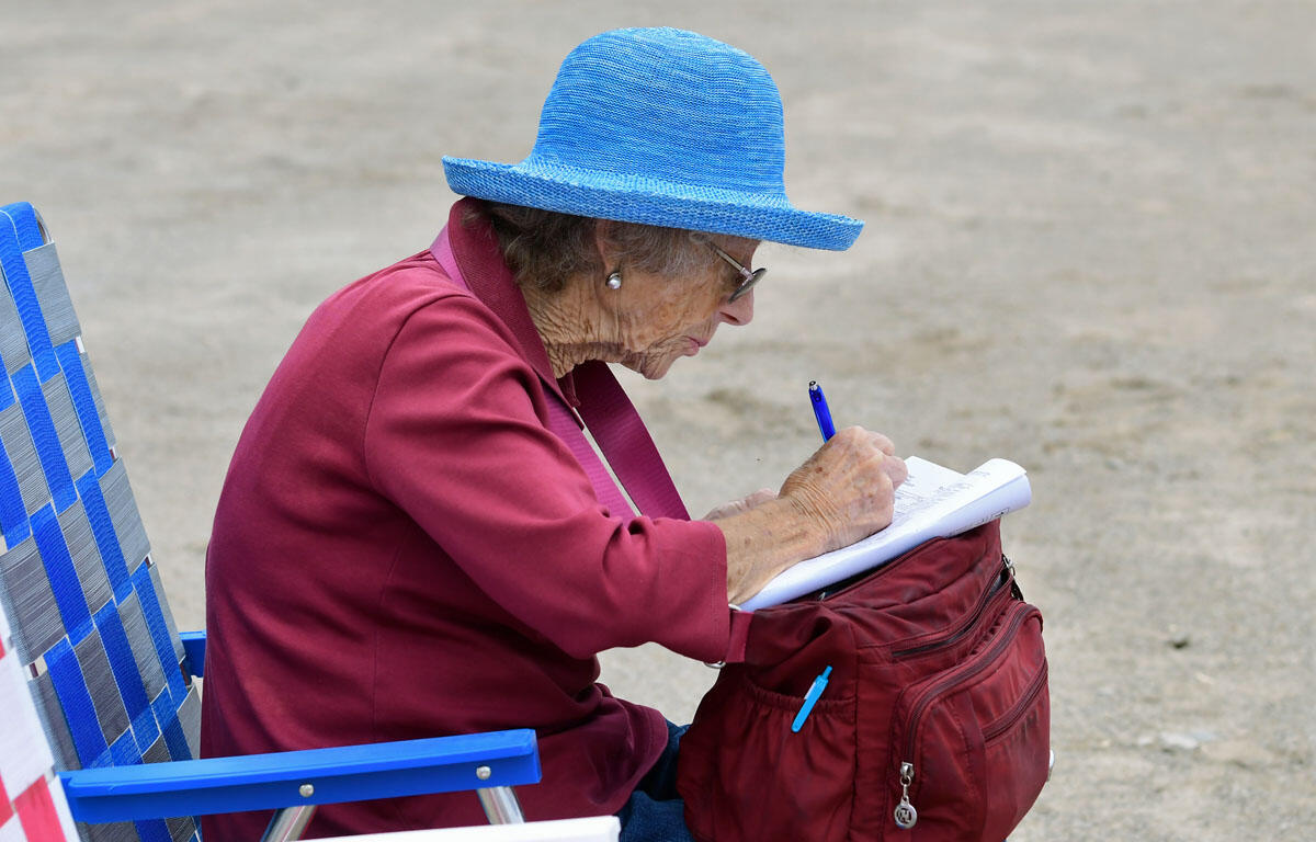 Elderly woman in a bright blue hat sits on a blue striped chair, writing in a notebook from a red backpack outdoors.
