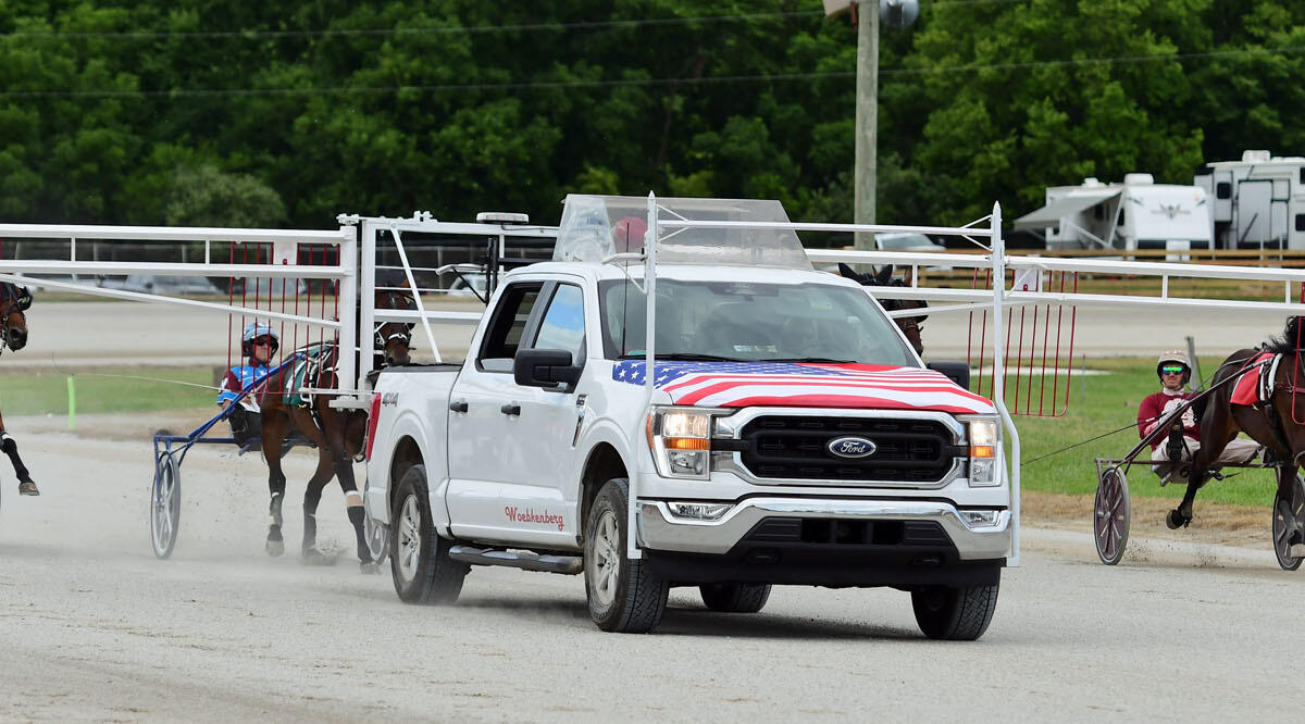 White pickup truck with American flag graphics leads harness racing sulkies on a dirt track.
