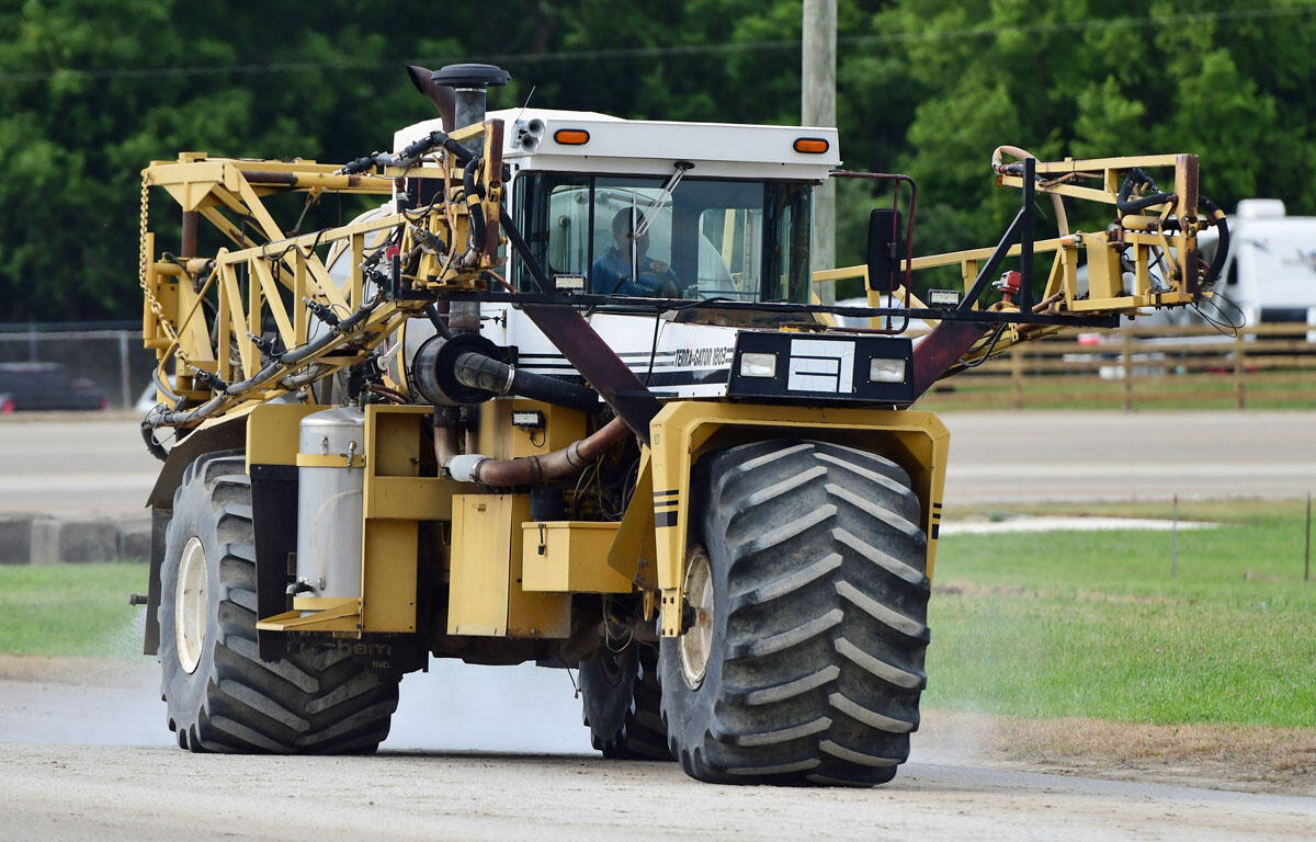 Large agricultural sprayer vehicle with extended arms driving on a dirt road, spraying field nearby.