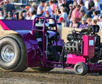 Pink drag racing car with a large rear tire on a dirt track; driver seated in open cockpit, crowd watching in the background.