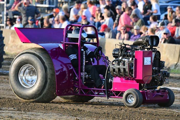 Pink drag racing car with a large rear tire on a dirt track; driver seated in open cockpit, crowd watching in the background.