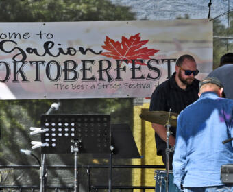 Stage at Galion Oktoberfest with a banner reading 'Welcome to Galion Oktoberfest' and a small band setup including a drummer and keyboard on the right side.